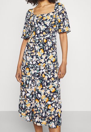 Frau trägt ein marineblaues Midikleid mit orange-, weißen und grünen Blumenmustern und kurzen Puffärmeln.