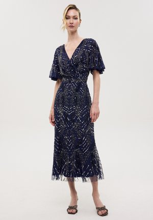 Vrouw draagt een marineblauwe midi-jurk met zilveren bloemenkralen, vliegende mouwen en V-hals, gecombineerd met open-toe hakken.