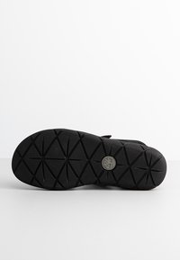 Suela de zapato negra con un patrón de textura triangular y un logotipo circular. Hecha de goma, presenta un diseño plano y antideslizante.