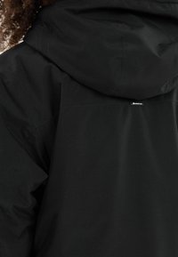 Schwarze Outdoor-Jacke mit Kapuze, strukturiertem Stoff und einem kleinen Markenetikett auf der Rückseite, mit nahtloser Verarbeitung, die an den Nähten sichtbar ist.