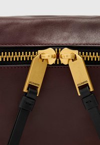 Detalj av burgunder skinnveske med svart glidelås og gullfarget utstyr, samt "ALLSAINTS" inngravert på glidelåsgrepene.