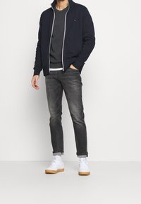 Granatowa bluza na zamek nałożona na ciemnoszary sweter z okrągłym dekoltem, zestawiona z obcisłymi szarymi dżinsami i białymi sneakersami z jasnobrązową podeszwą.