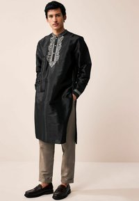 Black kurta made of silk with embroidered detailing on the neckline. Paired with light beige pants and dark brown loafers.