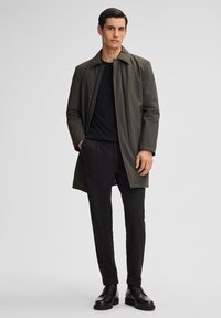 Cappotto verde oliva, con texture, dotato di colletto e tasche frontali, indossato sopra una camicia nera e pantaloni neri. Scarpe nere con lacci.