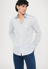 Camisa de rayas en blanco y azul con botones, presenta un cuello clásico, mangas largas y un logo sutil en el pecho. Tejido duradero.