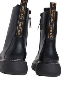 Stivali alla caviglia in pelle nera con suola in gomma spessa. Presentano una chiusura con cerniera e un'etichetta di colore nero e arancione con il marchio sul retro.