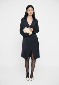 Rochie închisă la culoare, cu mâneci lungi și decolteu adânc în formă de V, având o tăietură asimetrică. Asortată cu o geantă de umăr într-o nuanță deschisă și colanți negri.