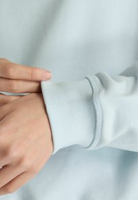 Felpa azzurro chiaro, polsino a costine, tessuto morbido, mano che sistema la manica, tessuto liscio, dettagli di cuciture sottili.