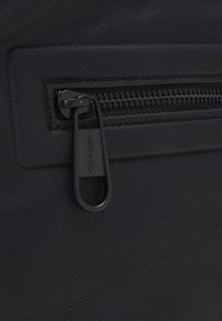 Dettaglio di una borsa in pelle nera con chiusura a zip, texture liscia e zip in metallo nero con il marchio "Calvin Klein" inciso.
