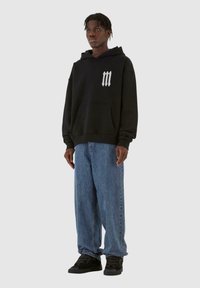 Felpa nera con cappuccio e tasca frontale, logo bianco sul petto, abbinata a jeans blu larghi e scarpe da ginnastica nere. Tessuto morbido.