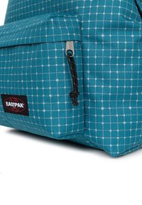 Zaino teal con un motivo a griglia, dotato di una tasca con cerniera e accessori neri. Realizzato in un tessuto testurizzato con un'etichetta del logo.