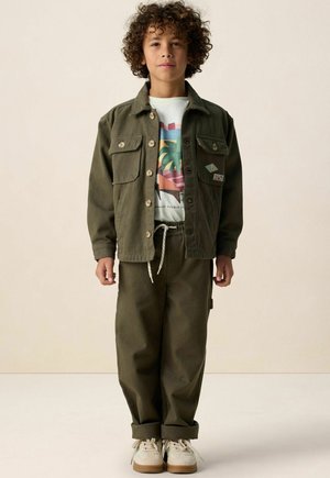Enfant aux cheveux bouclés portant une veste boutonnée vert olive et un pantalon assorti, un t-shirt graphique, et des baskets blanches, debout devant un fond uni.