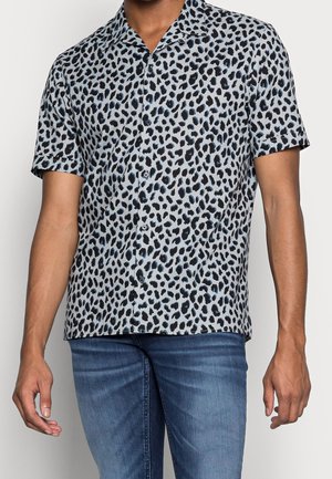 Mann trägt kurzärmeliges Hemd mit schwarz-blauem Leopardenmuster und blaue Jeans, steht vor weißem Hintergrund.