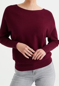 Person som bär en ribbad långärmad vinröd tröja och ljusgrå jeans, med händerna varsamt knäppta vid midjan, stående mot en vit bakgrund.