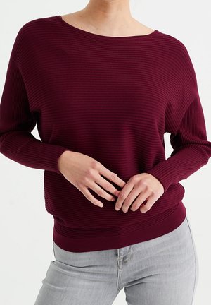 Persona che indossa un maglione a coste a maniche lunghe color amaranto e jeans grigi chiari, con le mani delicatamente incrociate in vita, in piedi su uno sfondo bianco.