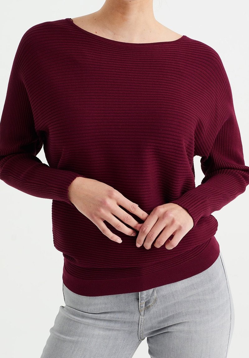 Person som bär en ribbad långärmad vinröd tröja och ljusgrå jeans, med händerna varsamt knäppta vid midjan, stående mot en vit bakgrund.