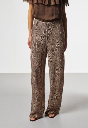 Vrouw draagt wijd uitlopende broek met hoge taille in een bruin-beige zebraprint, gecombineerd met een kortmouwige, doorzichtige bruine blouse en sandalen met open tenen.