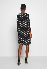 Vestido negro con lunares y mangas tres cuartos, dobladillo a la altura de la rodilla, escote redondo y detalle de botones en la parte posterior, combinado con tacones altos negros.