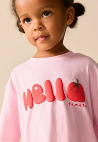 Světle růžové bavlněné tričko s výrazným červeným nápisem "HELLO" a obrázkem usmívajícího se rajčete. Kulatý výstřih, volný střih. Jemná textura.