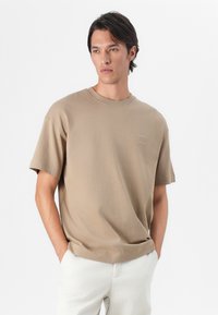Beige bomulds t-shirt med korte ærmer, rund halsudskæring og et subtilt brandtryk på brystet, sammen med lyse bukser.