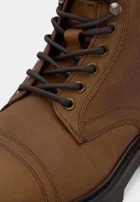 Bota de cuero marrón con cordones redondos oscuros, ojales de latón y costuras detalladas en la punta y los lados, mostrando "EST. 1990" grabado en la lengüeta.