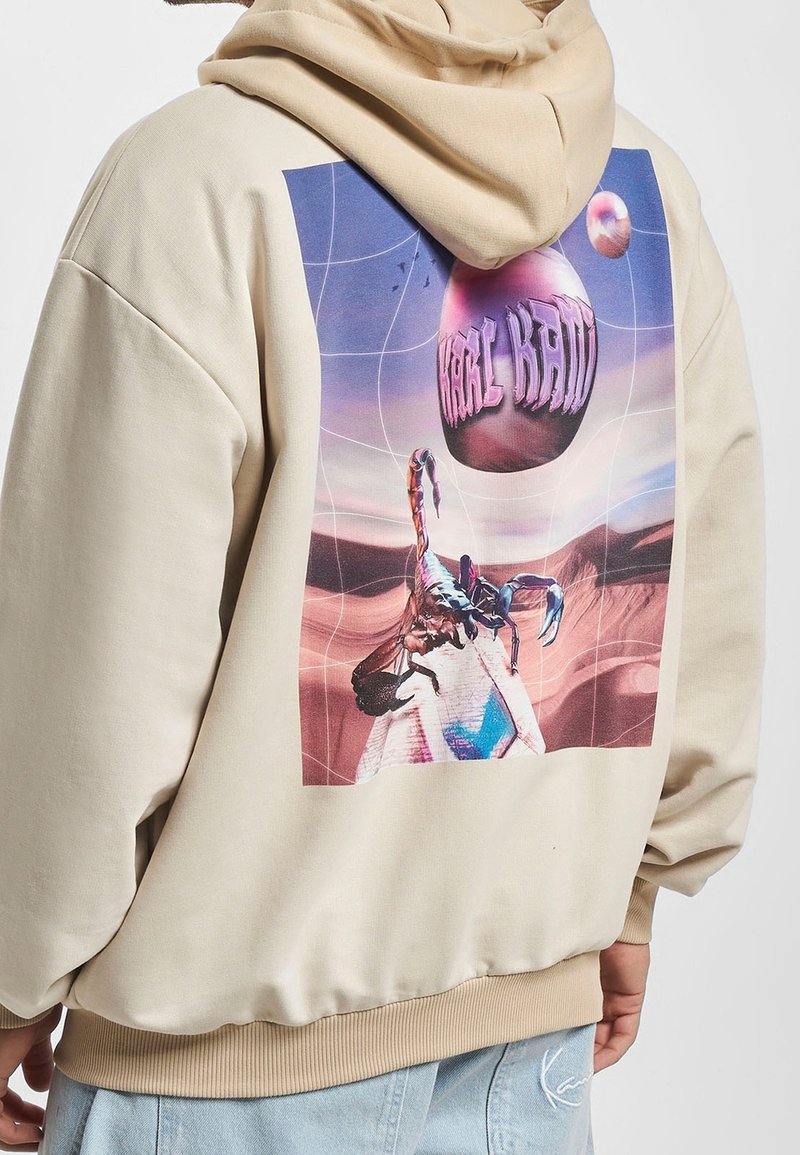 Sweat à capuche beige avec un graphisme coloré d’un scorpion sur une pyramide sous un orbe flottant avec un texte déformé sur un fond désertique surréaliste.
