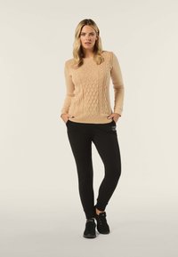 Beige Kabelstrickpullover mit langen Ärmeln, kombiniert mit enger schwarzen Jogginghose. Schwarze Sportschuhe vervollständigen das lässige Outfit.