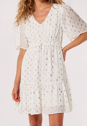 Femme portant une robe blanche à col en V avec un motif de feuilles dorées, taille à cordon, manches courtes et ourlet à volants, debout devant un fond uni.