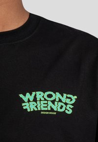 Černé bavlněné tričko s zeleným nápisem "WRONG FRIENDS" se zkresleným designem. "DESIGN HOUSE" je napsáno menším písmem níže.