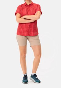 Camicia a maniche corte a quadri rossi con bottoni, abbinata a pantaloni corti beige. Modello che indossa scarpe da ginnastica color turchese con suola spessa.