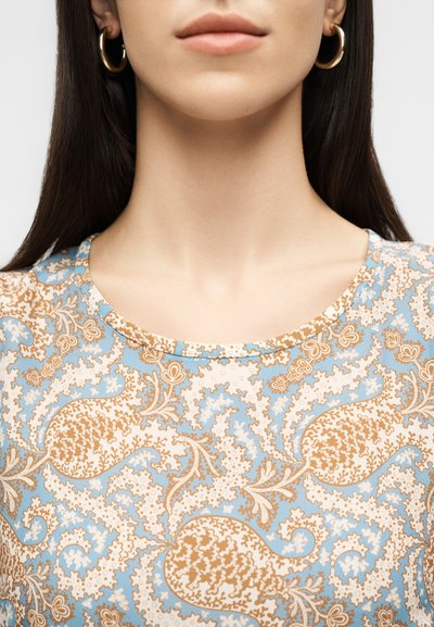 Γυναίκα με μακριά σκουρόχρωμα μαλλιά που φοράει χρυσούς κρίκους στα αυτιά και ανοιχτό μπλε τοπ με μπεζ και λευκό περίτεχνο λουλουδάτο μοτίβο.