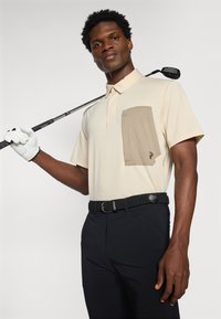 Camisa polo bege com um bolso castanho, mangas curtas e colarinho; combinada com calças pretas e um cinto trançado; segurando um taco de golfe.