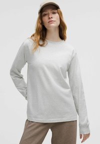 Lys grå langærmet skjorte lavet af blødt stof med rund halsudskæring og lige snit; parret med brune teksturerede bukser og en beige kasket.