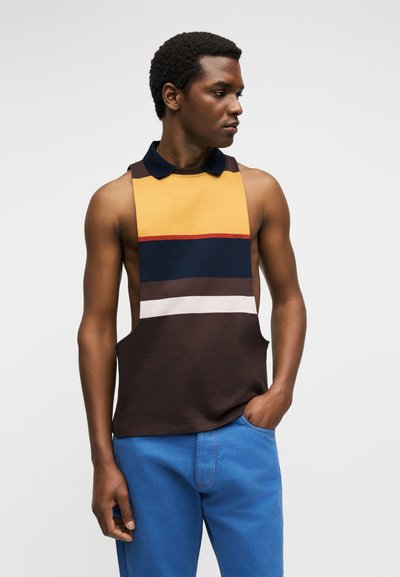 Άνδρας που φοράει αμάνικο πόλο μπλουζάκι με κίτρινες, μπλε σκούρες, καφέ και λευκές ρίγες και μπλε παντελόνι, κοιτάζει στο πλάι με το χέρι στην τσέπη.