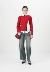 Jongeman staat, gekleed in een rood asymmetrisch knoopshirt, witte blouse met zwarte stropdas, vervaagde jeans, zwarte hakken, en houdt een kleine bordeauxkleurige tas vast.