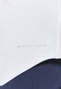 Bílé sportovní tričko Omni-Wick s hladkou texturou, které má jemné logo a kontrastní šití. Zobrazeno nad tmavě modrou látkou.