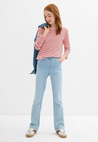 Maglietta a righe rossa e bianca a maniche lunghe, pantaloni in denim azzurro chiaro a zampa di elefante e sneakers bianche con dettagli neri. Giacca di denim appoggiata sulla spalla.