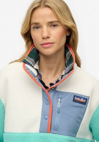 Pull en polaire couleur crème, aqua et bleu avec une poche poitrine zippée, un col à carreaux et des finitions orange. Tissu texturé sur l'ensemble.