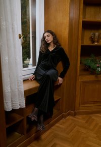 Svart sammetsklänning med långa ärmar och detalj med knytband. Bärs med genomskinliga svarta strumpor och prickiga klackskor. Interiör med träpaneler.