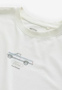 Valkoinen lyhythihainen paita, jossa on sininen grafiikka Ford-pickupista ja logosta, rento pyöreä kaula-aukko ja pehmeä puuvillakangas.