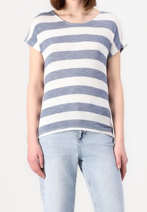 Vrouw met een blauw-wit horizontaal gestreept T-shirt met korte mouwen en lichtblauwe spijkerbroek, staande tegen een effen achtergrond.