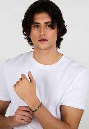 Bracciale in tono argento indossato su un polso, caratterizzato da un design a catena. Modello con t-shirt bianca semplice e capelli castano scuri, lunghi fino a metà lunghezza e mossi.