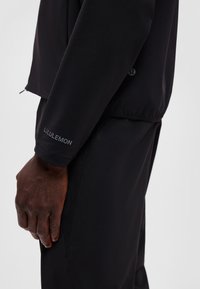 Zbliżenie osoby noszącej czarną kurtkę i spodnie Lululemon, pokazujące lewą rękę i bok z logo Lululemon na rękawie kurtki.