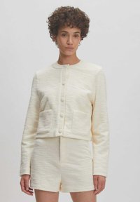 Crèmekleurige textuurjacket met ronde halslijn en knoopsluiting, gecombineerd met bijpassende shorts. Opvallende voorzakken en een getailleerde pasvorm.