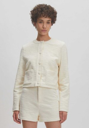 Cream textured jacket with round neckline and button closure, paired with matching shorts. Notable front pockets and tailored fit.