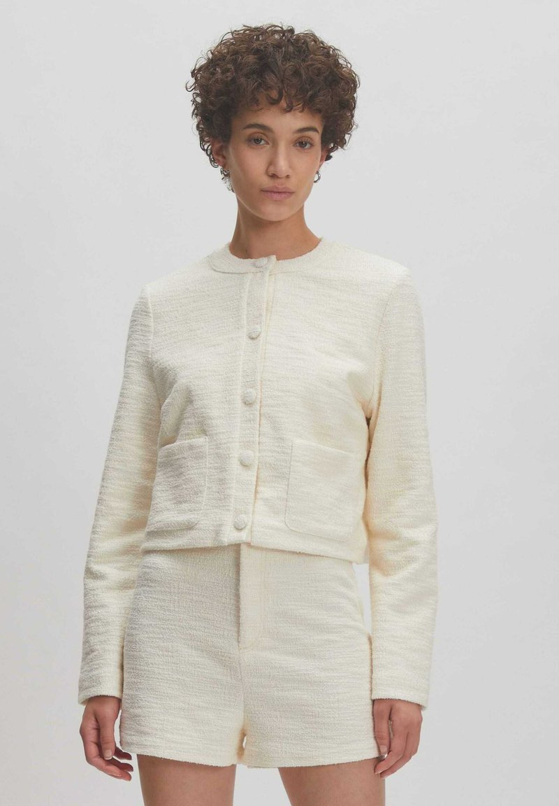 Crèmekleurige textuurjacket met ronde halslijn en knoopsluiting, gecombineerd met bijpassende shorts. Opvallende voorzakken en een getailleerde pasvorm.