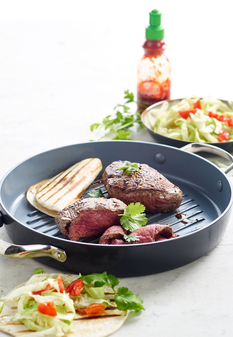 Trozos de carne asada y pan plano en una sartén, con un toque de cilantro, ensalada con lechuga y tomates, y una botella de salsa picante al fondo.