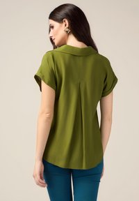 Olijfgroene blouse met korte mouwen en een kraag, met een achterste plooid ontwerp en een gladde stofstructuur. Gecombineerd met blauwe broek.
