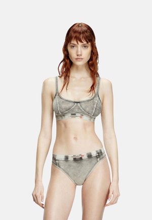 Donna con lunghi capelli rossi sta in piedi di fronte, indossando reggiseno e slip grigi coordinati con stampa denim su uno sfondo chiaro e semplice.