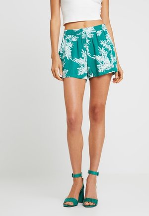 Femme portant un short vert à motif tropical et des sandales vertes à talons hauts assorties, debout sur un fond uni.